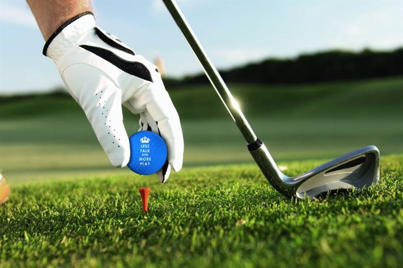 Hand im Golfhandschuh setzt einen blauen Keep-Calm-Golfball mit Aufdruck auf ein rotes Tee, daneben ein Eisen