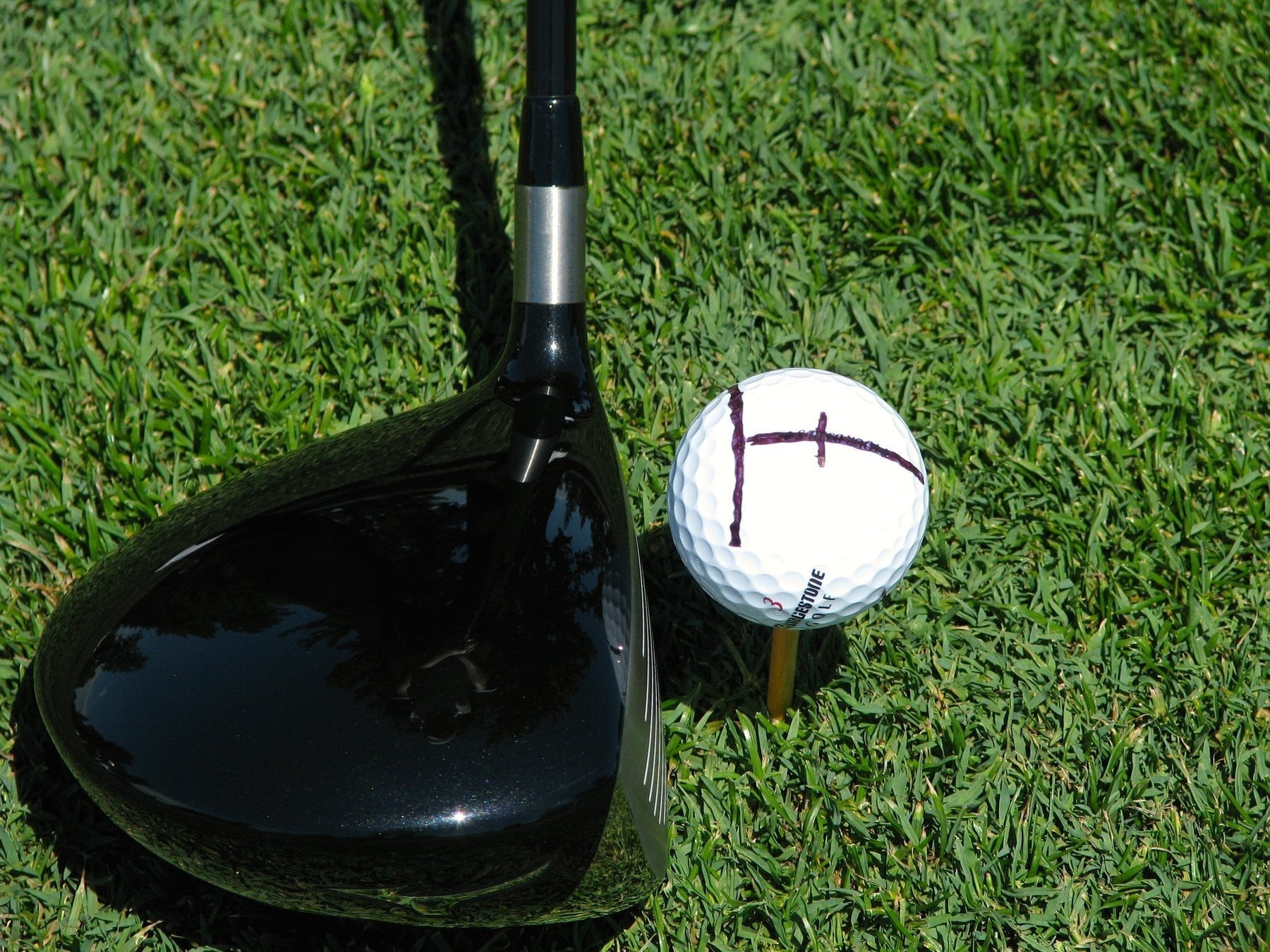 Golfball mit aufgezeichneter Ziellinie auf dem Tee, daneben ein schwarzer Driver im Ansprechen